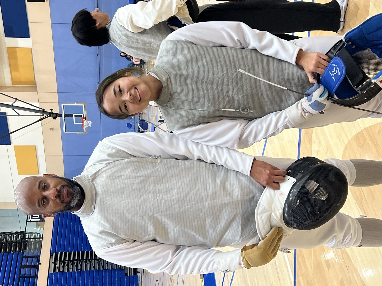 A fencing coach and a young competitor in full white gear smiling on the gym floor